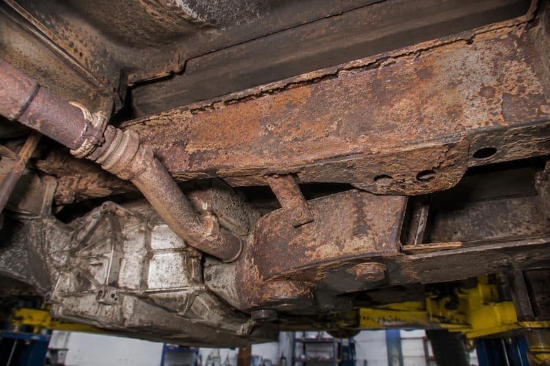 Scaled and rusting rear chassis rail running above the gearbox on a VW T25, with the exhaust downpipe and rear crossmember in shot.