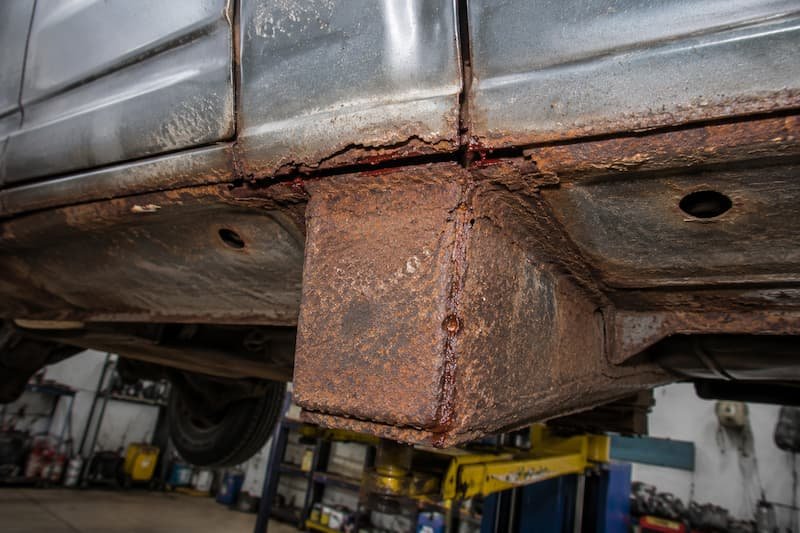 Active corrosion in the corner joint where the outrigger meets the sill on a VW T25, with the seam opening up and factory drain holes either side.