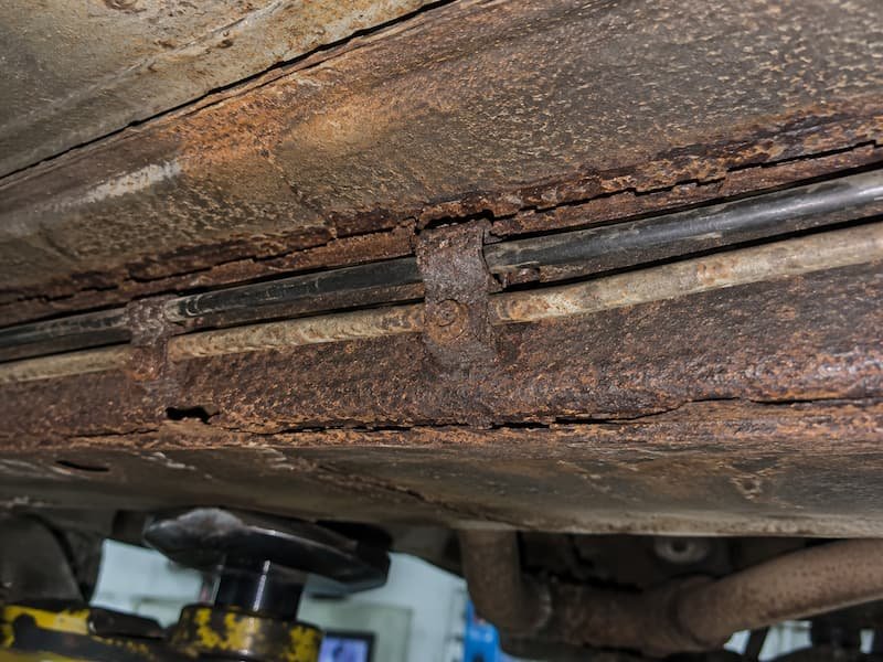 Brake and fuel lines clipped to a heavily corroded sill panel on a VW T25, showing the P-clip holding the pipe against the rusted steel.