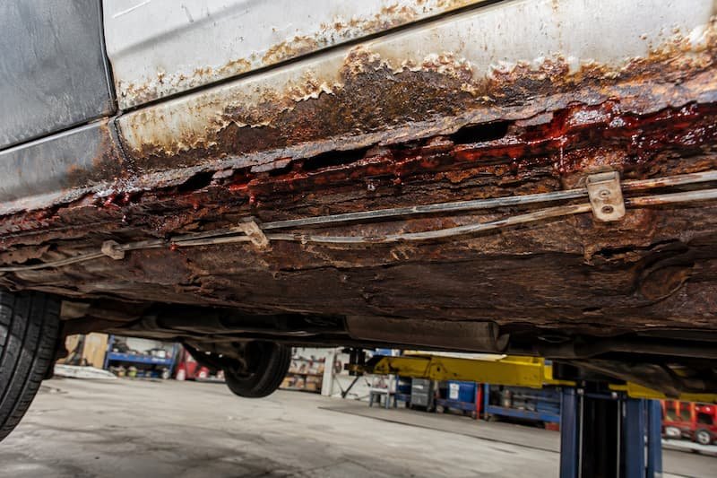 Heavy laminating rust and bright red active corrosion along the nearside sill to floor seam of a VW T25, with brake and fuel lines clipped to the corroded section.