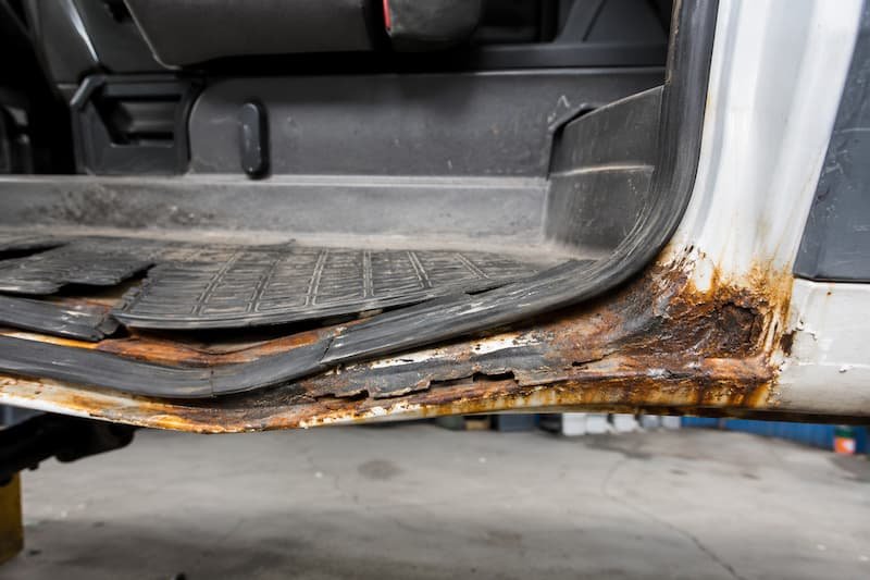 Rust blooming along the front edge of a VW T25 cab step, rubber tread lifting from the metal, and bubbling at the base of the B-post where it meets the sill.