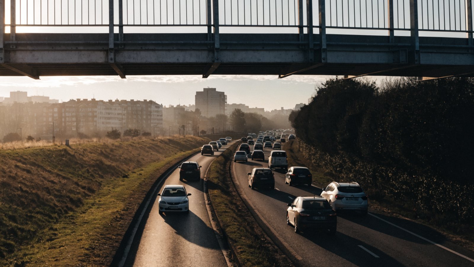 Morning commuter traffic in both directions on a dual carriageway at the edge of a city in low winter sun