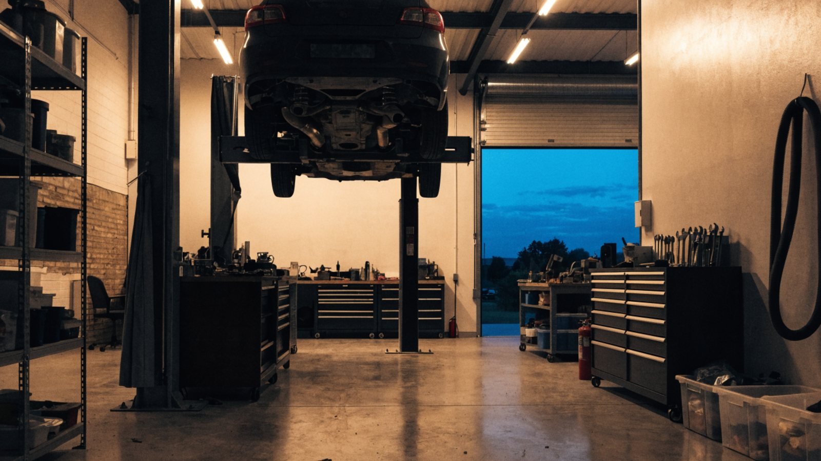 Garage workshop in the early evening with warm lights on, a car raised on a two post lift and a dark blue sky through the open door