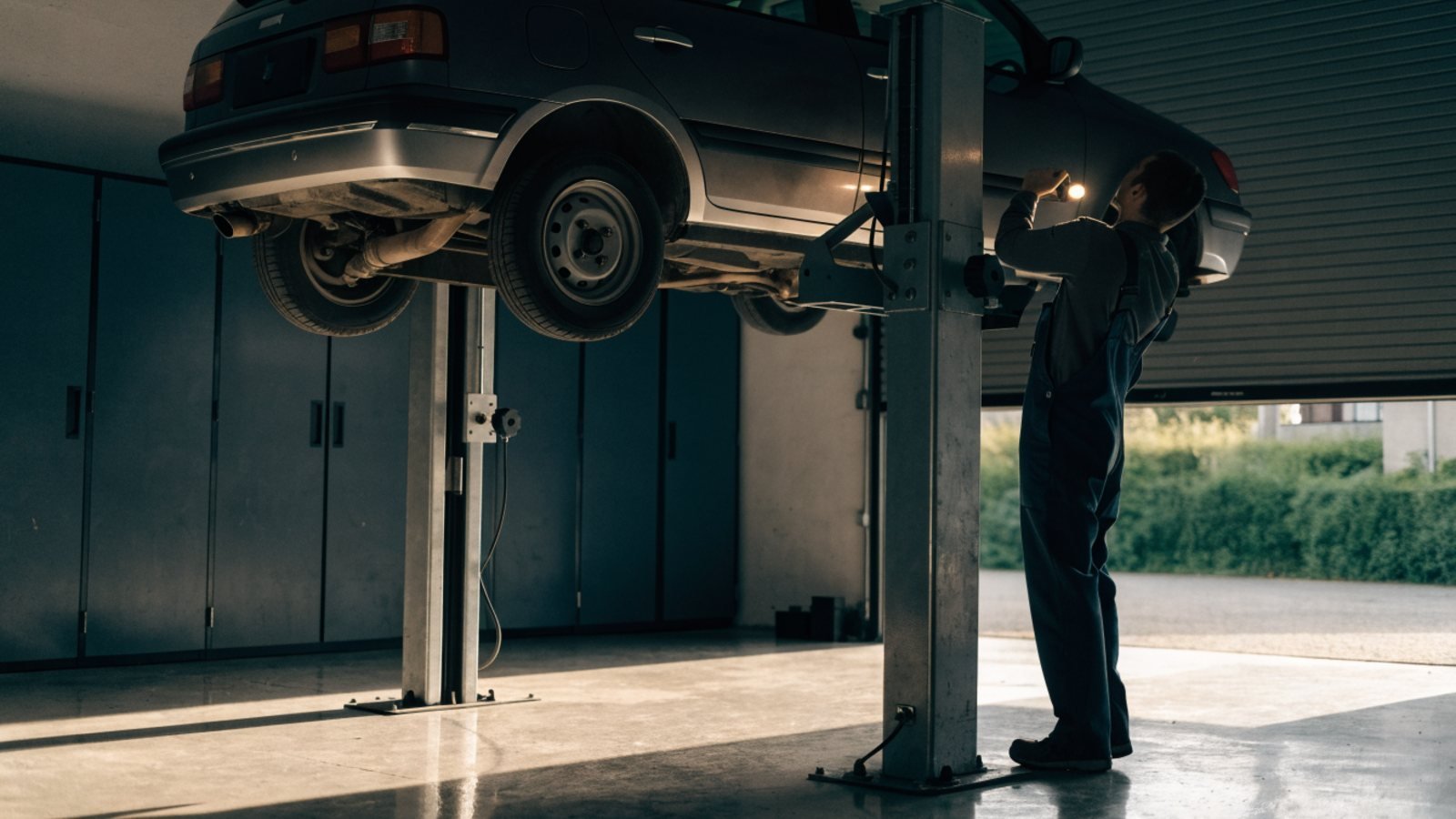 Mechanic in dark overalls inspecting the underside of a hatchback raised on a two post lift in an independent garage