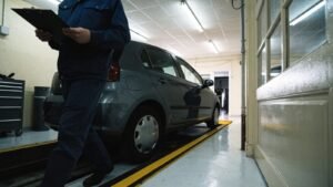 MOT tester in navy workwear with a clipboard walking beside a grey hatchback parked over an inspection pit in a garage
