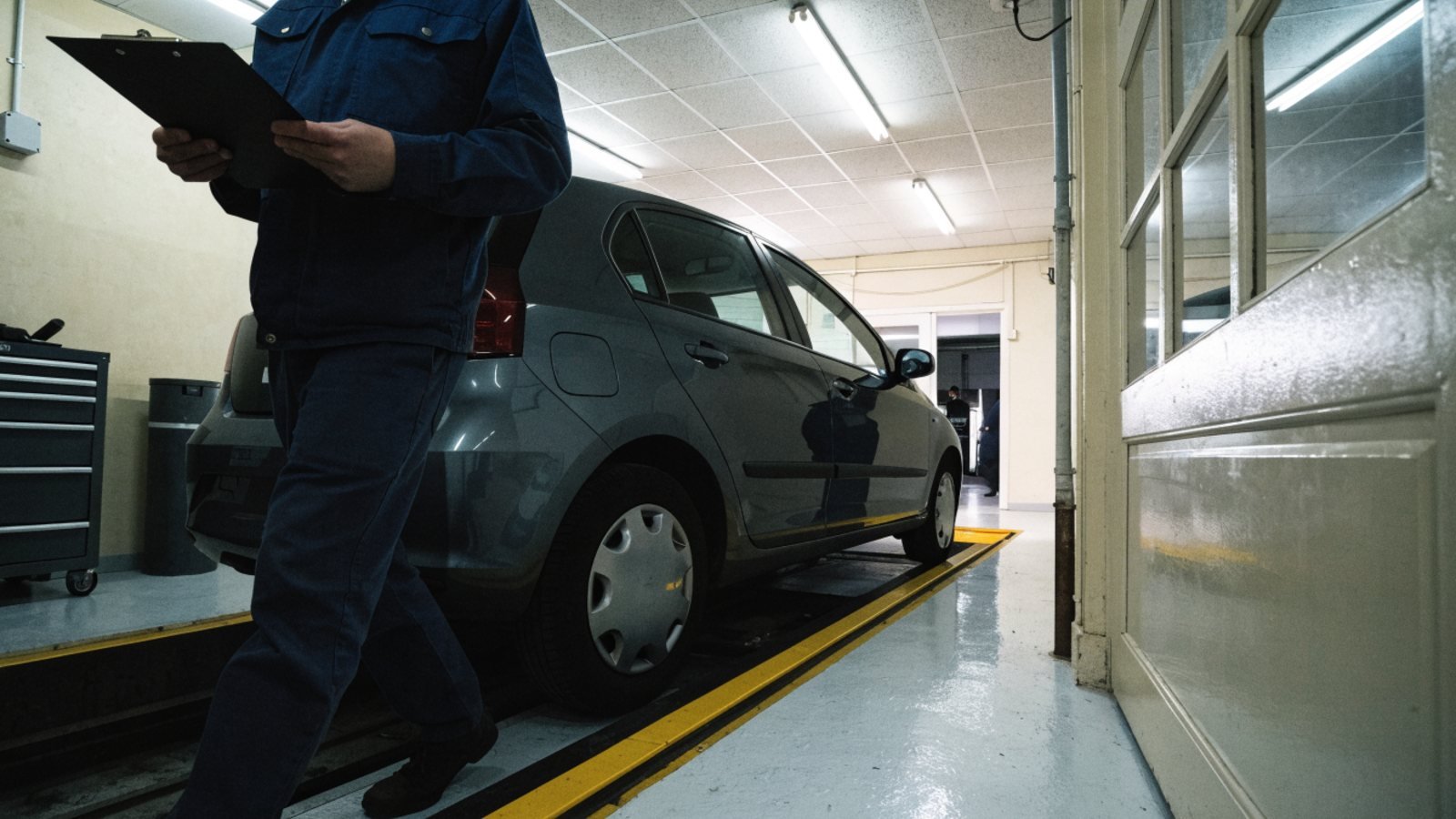 MOT tester in navy workwear with a clipboard walking beside a grey hatchback parked over an inspection pit in a garage