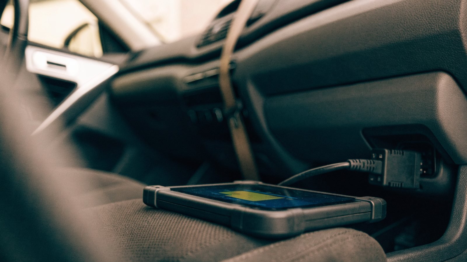 Diagnostic scan tool cable plugged into the port under a car dashboard with a tablet resting on the seat showing a live data graph