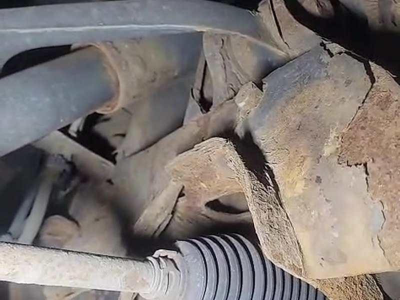 Thick flaking rust on the subframe mounting of an Iveco van next to the steering rack gaiter, seen from underneath on a workshop lift.