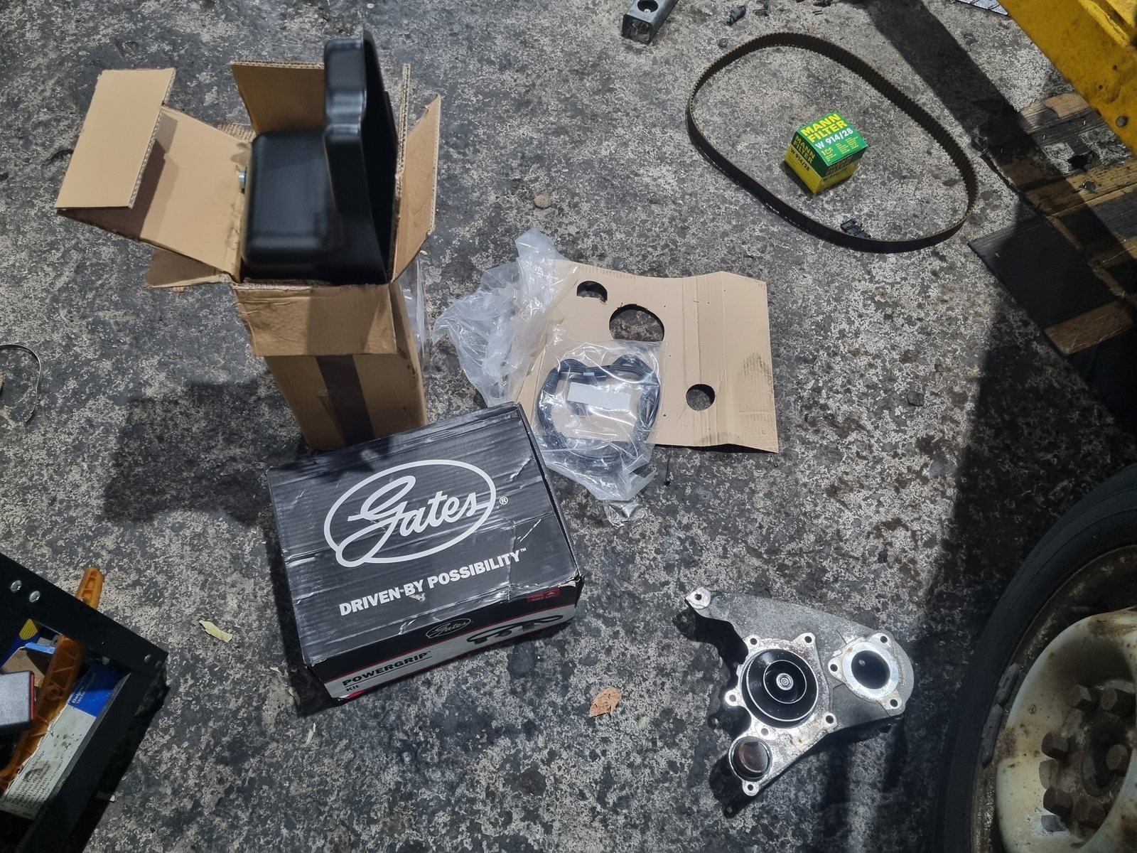 New timing belt kit box, replacement steel engine sump and gasket, a new oil filter, the old timing belt and the old water pump in its housing laid out on the workshop floor at CF3 MOT and Service Centre.