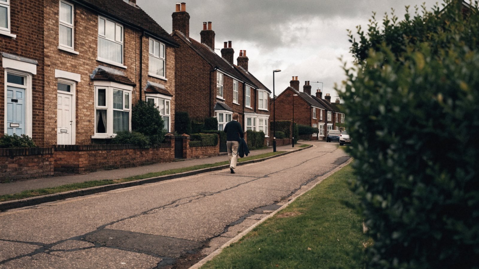 Person walking along a pavement past brick semi-detached houses in a suburban street on an overcast afternoon