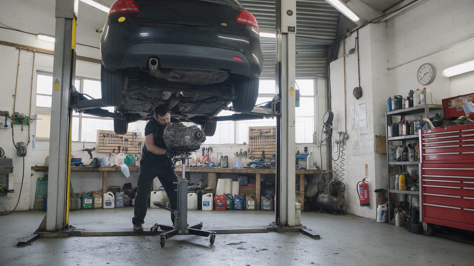 Technician guiding a gearbox onto a transmission jack under a hatchback on a two post ramp